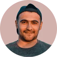Young man with a trimmed beard wearing a backward dark baseball cap and gray shirt, smiling slightly, against a solid muted pink background.