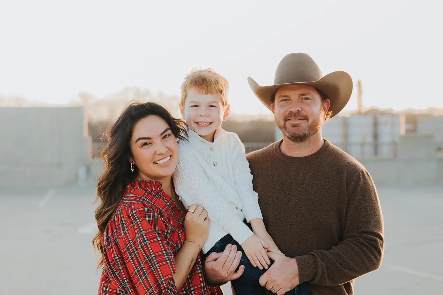 Smiling family of three with woman in red plaid, man with cowboy hat, holding a young boy in white sweater.