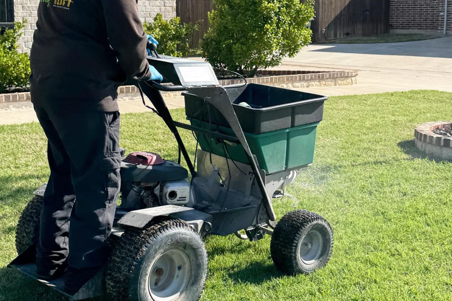 Person standing on a riding lawn spreader machine, wearing black pants and blue gloves, applying material to a green grassy lawn near a fire pit and bushes.