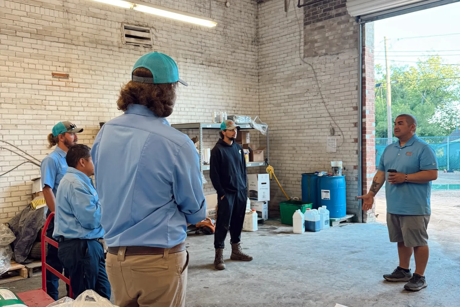 Five men standing inside a warehouse or workshop with brick walls, engaging in a discussion; three men wear light blue shirts and caps, one wears a black hoodie and boots, and one in a blue polo holds a cup while speaking near an open garage door.