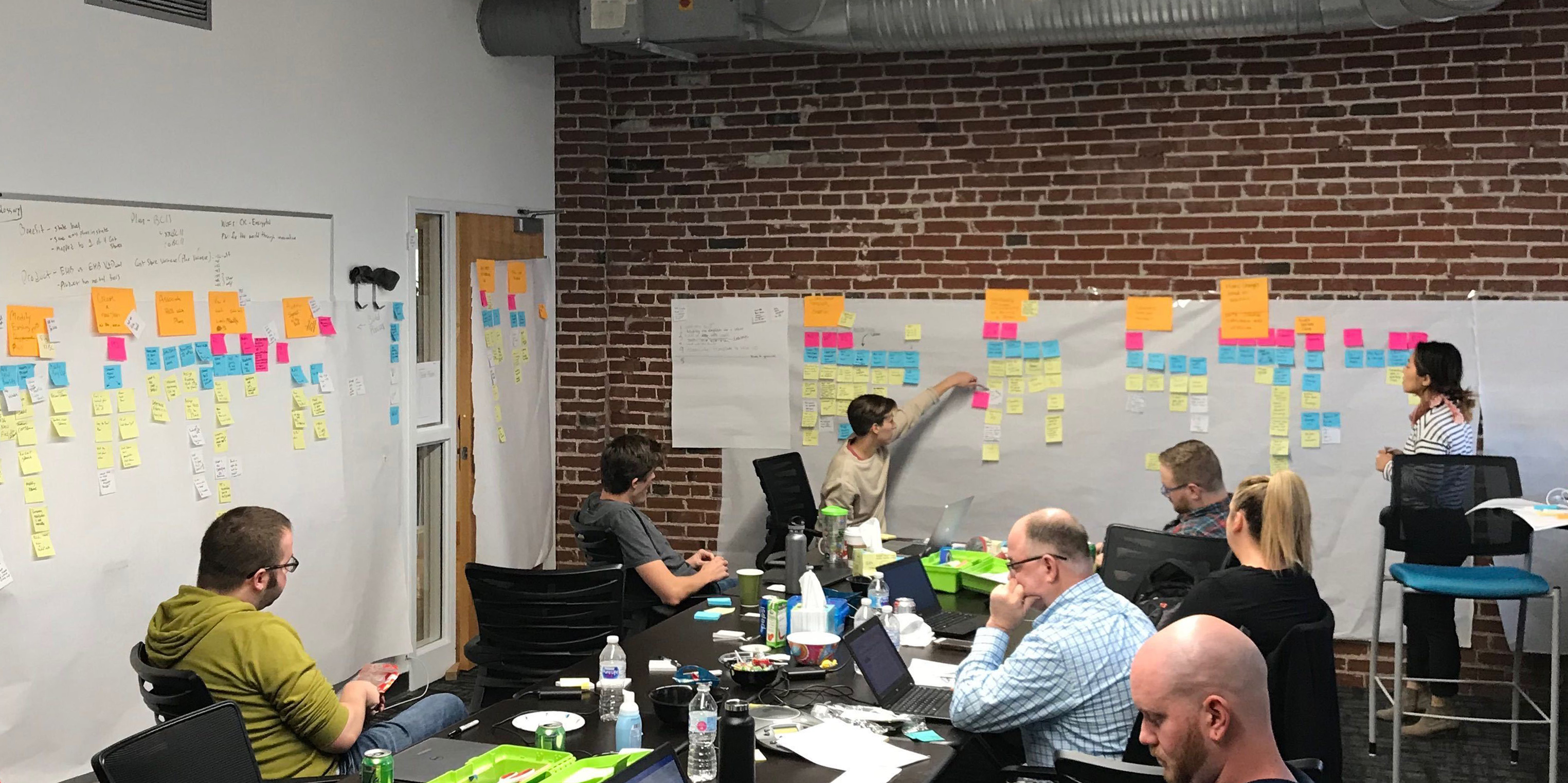 Group of people in a workshop sitting around a table with laptops, engaged in a story mapping session using sticky notes on large white sheets taped to a brick wall.