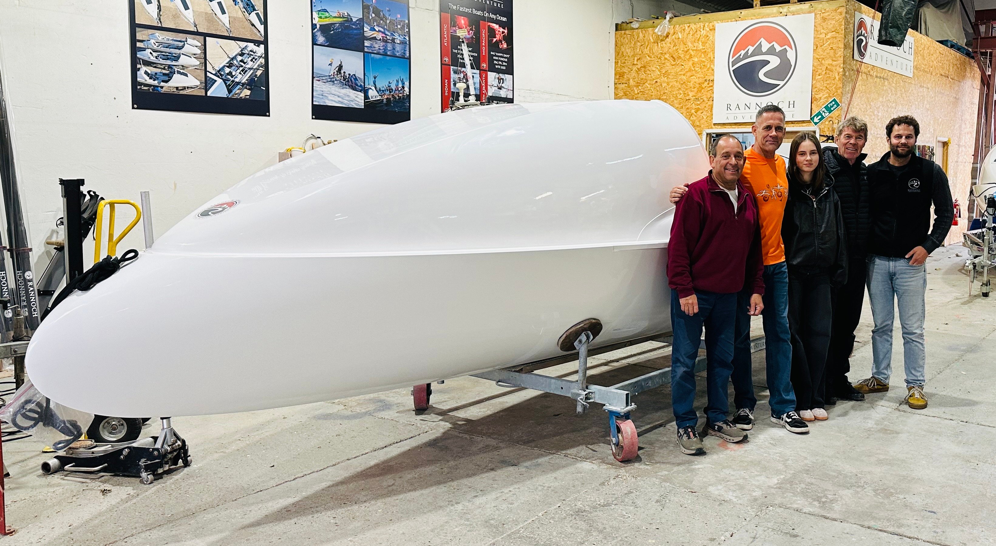 Five people standing beside a large, white boat hull in a workshop.