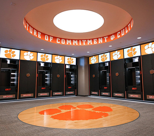 Clemson University football locker room with black lockers, orange paw prints, and a circular ceiling light displaying 'Culture of Commitment.'