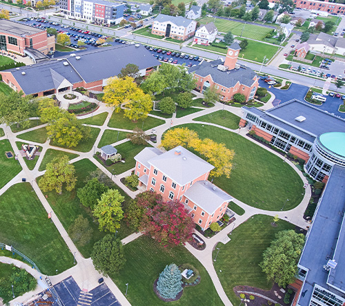 Aerial view of a college campus featuring multiple brick buildings, pathways, green lawns, and trees with autumn foliage.