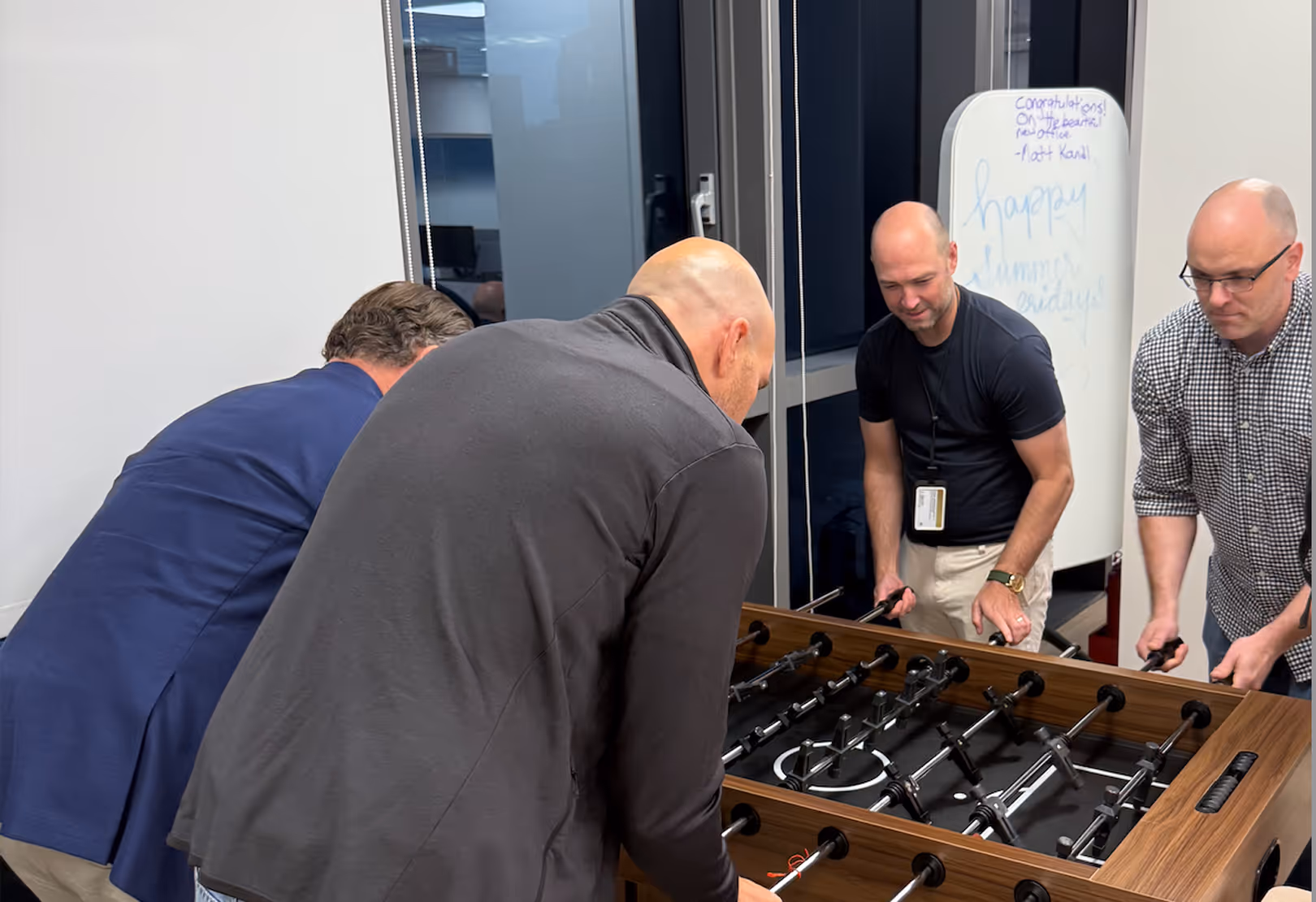 Four men engaged in a foosball game indoors, two men on each side of the table.