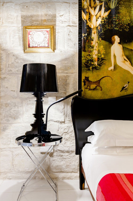 Orange basket with sunglasses on the bed implying a day at the beach beckons. The bed is unmade. Bed canopy detail with religious scene in background, a black Kartell Bourgie Table lamp on bedside table to the left of the picture. Malta holiday accommodation.