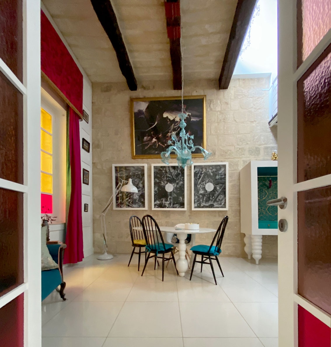The dining room as seen through open doors with coloured glass. The figure of eight shaped marble table on three voluptuous legs surrounded by black chairs under a Murano glass chandelier. Very high ceiling with wooden beams. Large framed artworks on the wall. Part of the white bespoke open wardrobe with same style voluptuous legs and with a gold and green interior. Malta boutique vacation rental.   