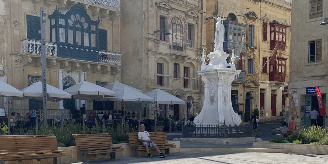 Birgu's main square with umbrellas and statue on large pedestal, man on bench and houses with elaborate balconies surrounding the square