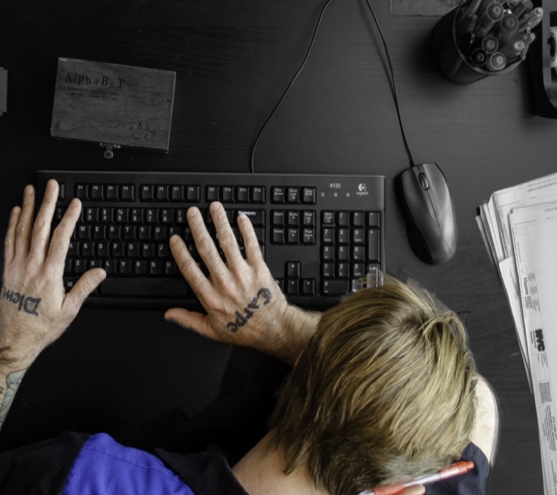 Man with tattoos laying his head on his desk in frustration, surrounded by inspection paperwork.