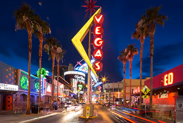 Night scene of a busy Downtown Las Vegas street with a large illuminated 'VEGAS' sign and palm trees.