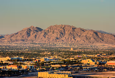 Cityscape with buildings and streets in front of large, Sunrise Mountain rocky mountains under a clear sky at sunset.
