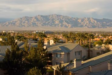 Las Vegas residential neighborhood with houses and palm trees in the foreground and mountain range in the background under a cloudy sky.