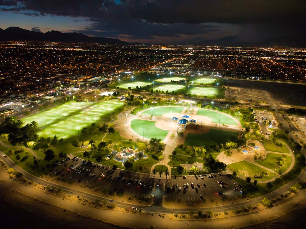 Desert Breeze Park. Curved paved walkway through a park with green grass, trees, and blue-roofed picnic shelters under a clear sky.
