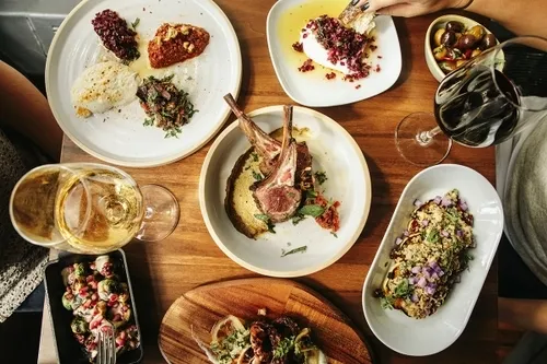 Overhead view of a wooden table with plates of gourmet dishes including lamb chops, creamy spreads with toppings, roasted vegetables, grilled octopus, olives, and two glasses of wine, one red and one white.