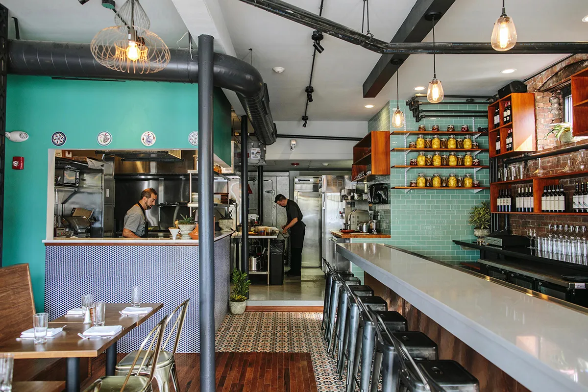 Modern restaurant interior with a long white counter, metal stools, turquoise tiled wall, shelves with jars and wine bottles, and two chefs working in the kitchen.