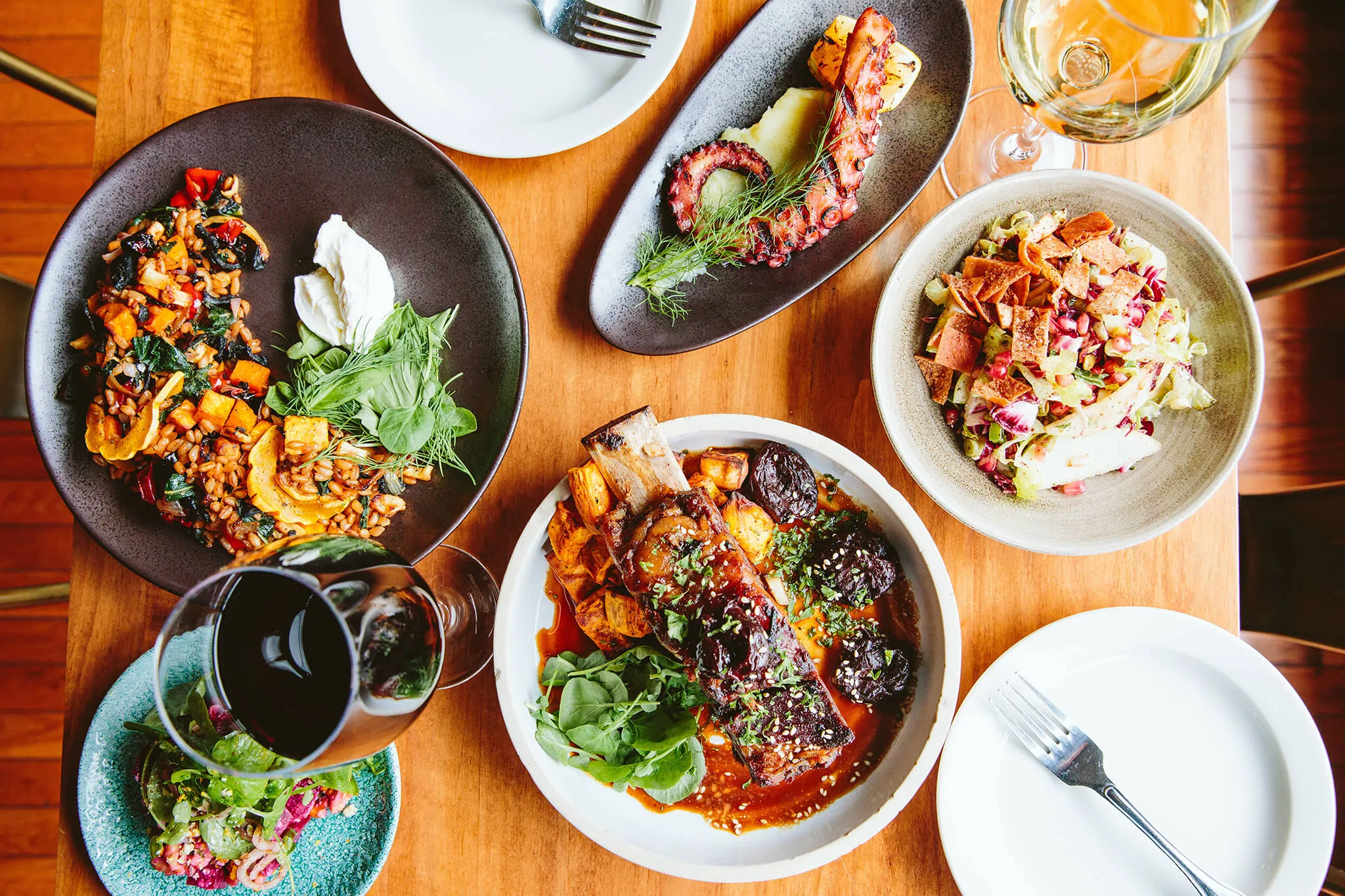 Top-down view of a wooden table set with various dishes including roasted meat with sauce, grilled octopus, vegetable salad, and two glasses of red and white wine.