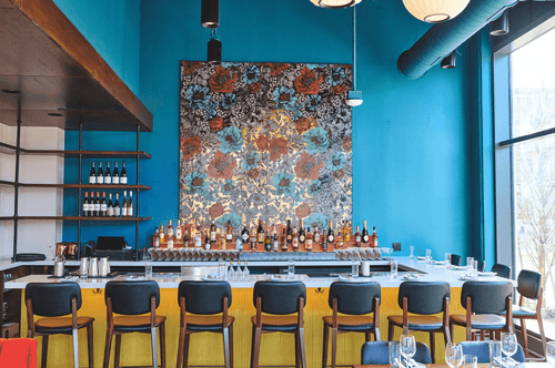 Modern bar with yellow paneling, wooden bar stools, blue walls, large floral artwork, and bottles of liquor on the countertop.