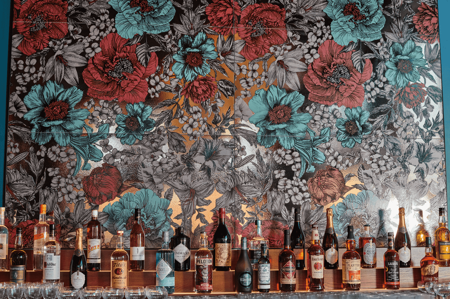 Bar shelf with various bottles of liquor in front of a large colorful floral mural featuring red, turquoise, and white flowers.