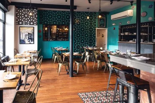 Cozy restaurant interior with wooden floors, turquoise walls featuring black geometric patterns, metal chairs, wooden tables set with glasses and napkins, and a bar area with stools.