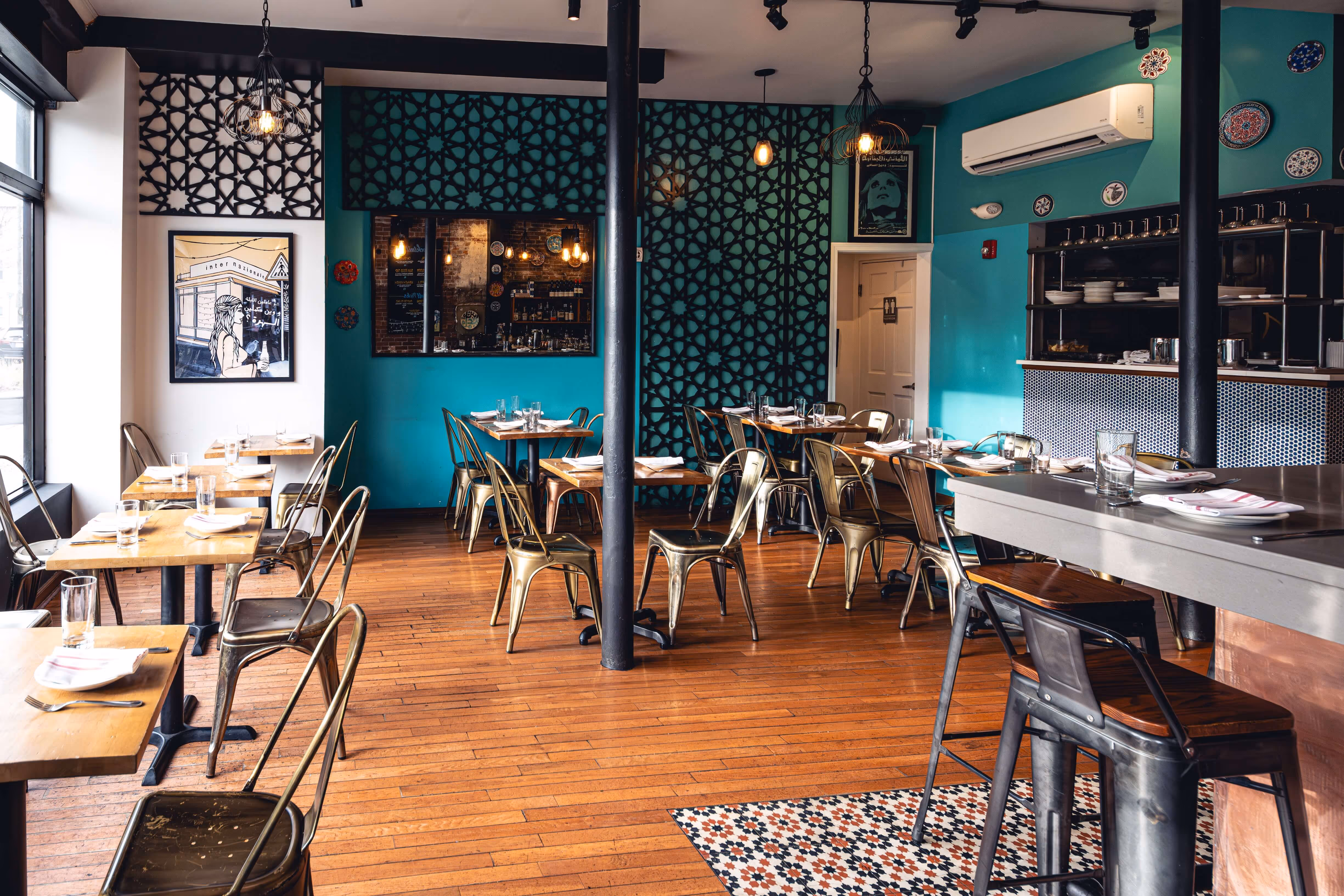 Cozy restaurant interior with wooden floors, turquoise walls featuring black geometric patterns, metal chairs, wooden tables set with glasses and napkins, and a bar area with stools.