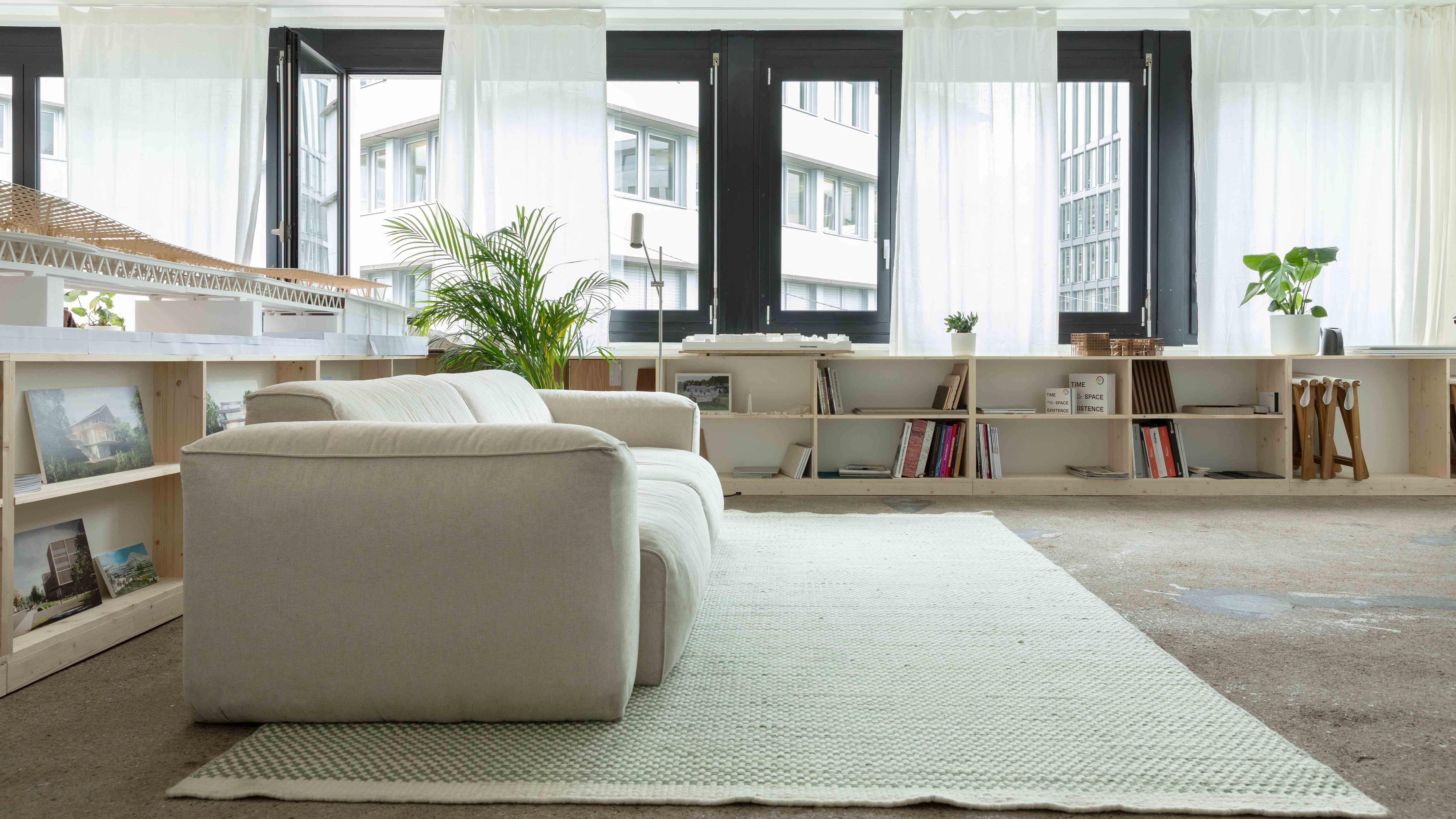 Modern office lounge with beige sofa, potted plants, wooden shelves, and large windows with white curtains.