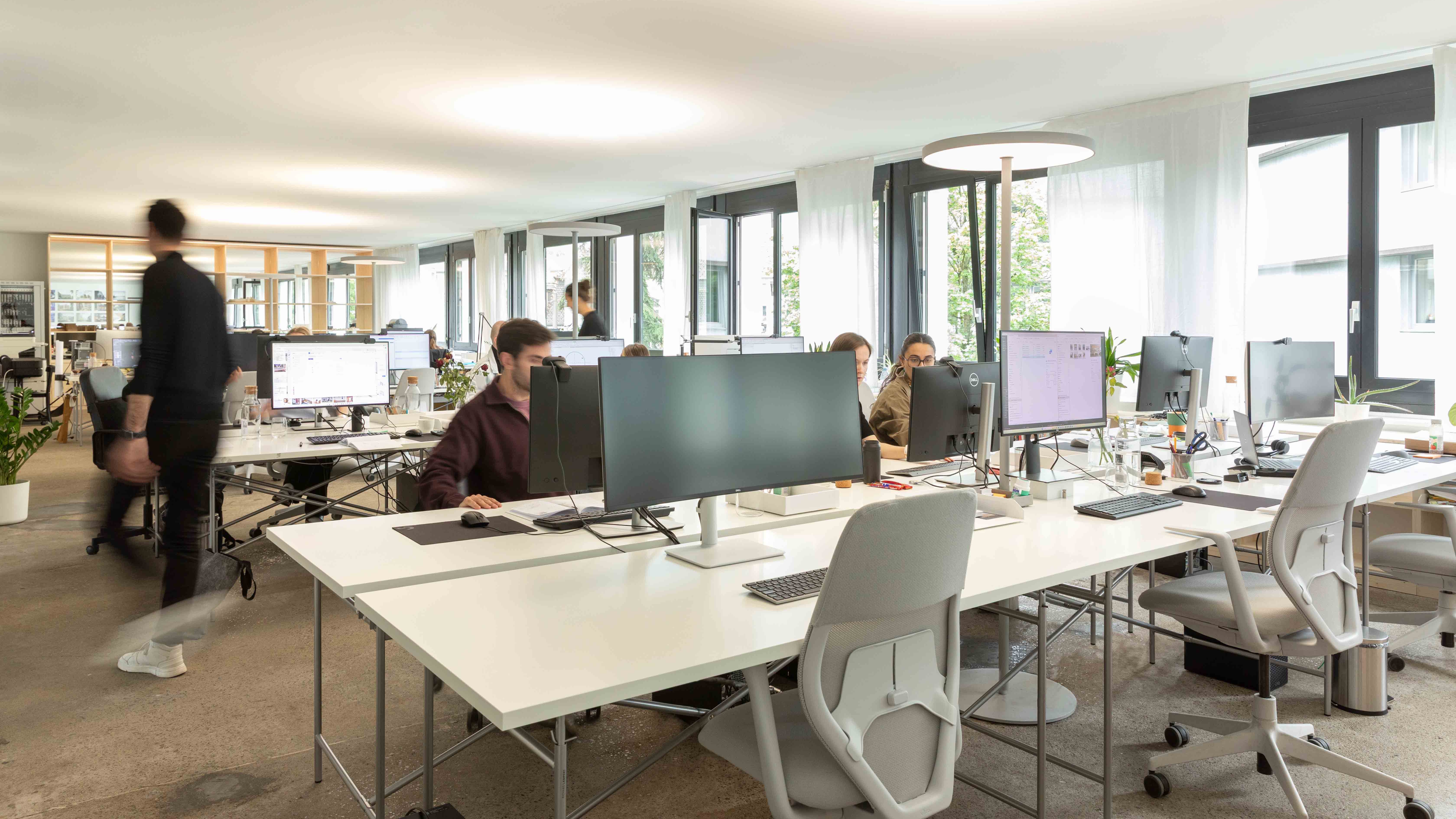 Modern, bright office space with workers at desks using multiple monitors and natural light from large windows.