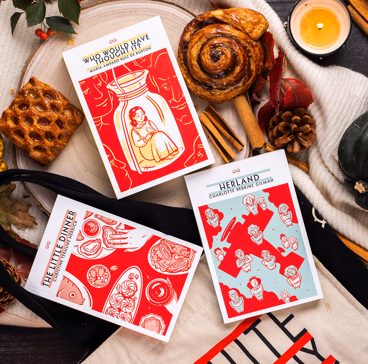 A spread of books from Quite Literally Books. There are three books, "Who Would've Thought It?" "The Little Dinner" and "Herland" The books are showcased alongside decorative pastries and candles.  