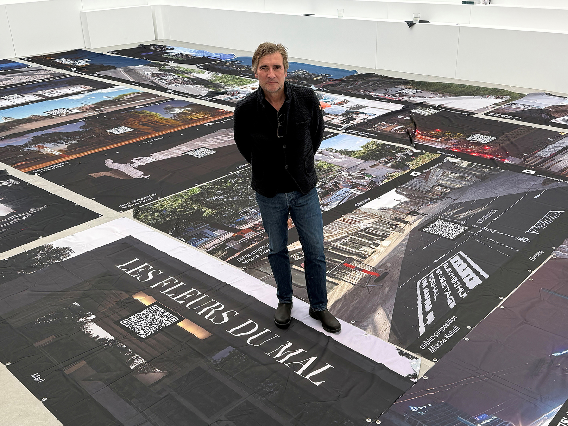 Künstler Mischa Kuball im Lichthof des Baukunstarchivs NRW vor dem Aufbau der „Bauzäune“. Bannerpräsentation mit zentralem Fotomotiv + QR-Code auf eine frühere public preposition in einer internationalen Großstadt verweisend., © VG Bild-Kunst, Bonn 2026, Foto: Christof Rose / Architektenkammer NRW