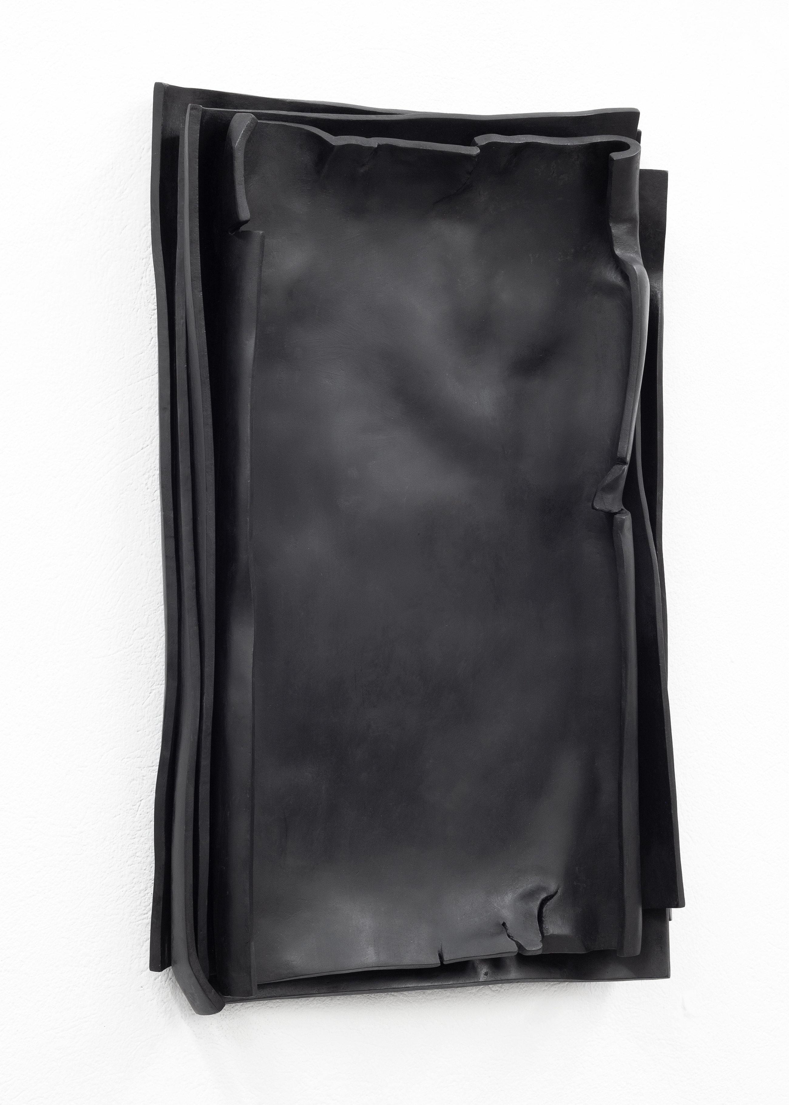 The photo shows a side view of the black epoxy resin sculpture Besser Vertikal 11. The artwork is installed hanging on a white wall. This work by sculptor Erik Andersen consists of several sheets resembling paper stacked on top of each other, which, defying the laws of gravity, appear to be loosely stacked vertically.