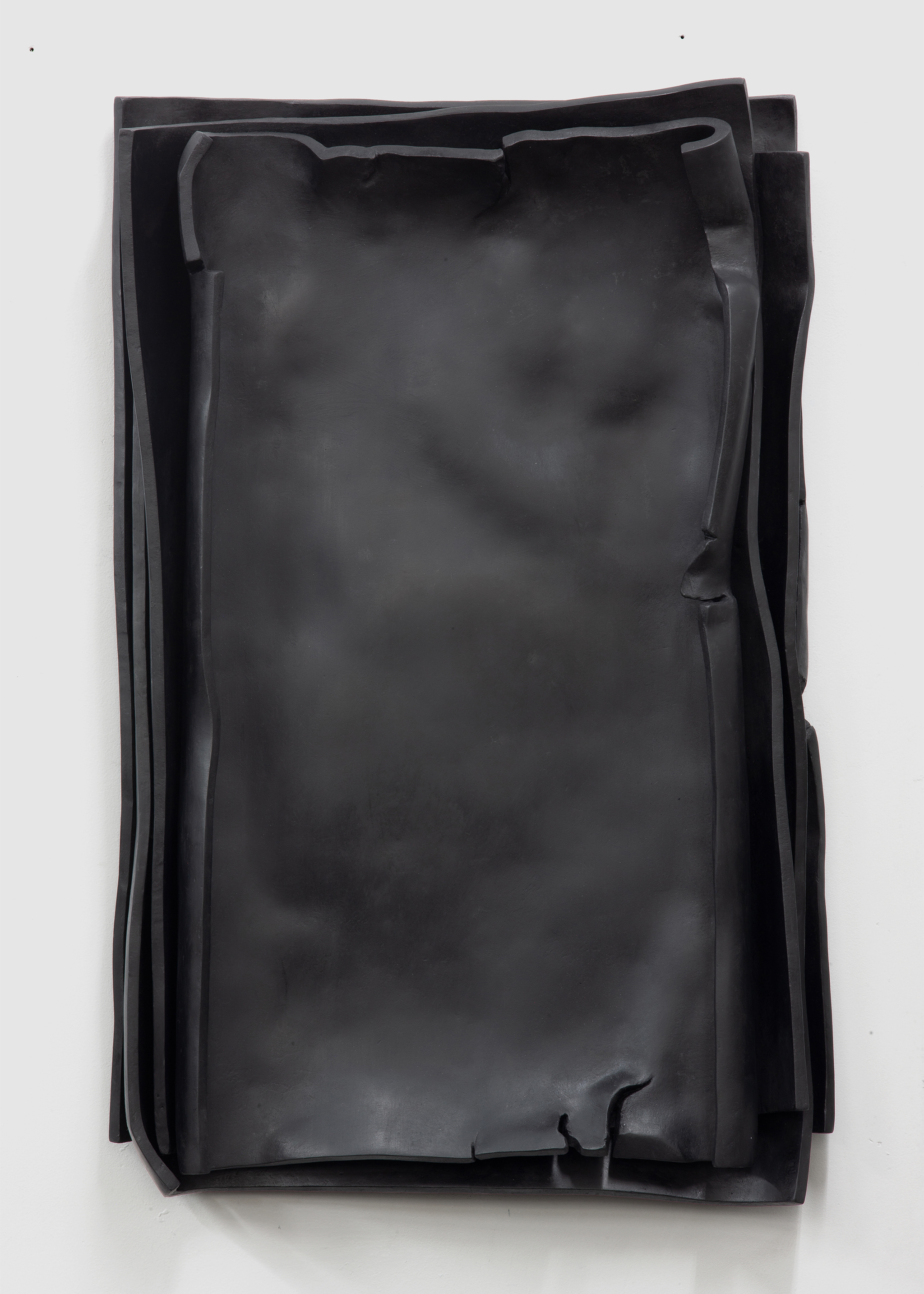 Front view of a black sculpture made of epoxy resin hanging on a white wall. The work by sculptor Erik Andersen appears to consist of a loosely stacked pile of sheets of paper. 