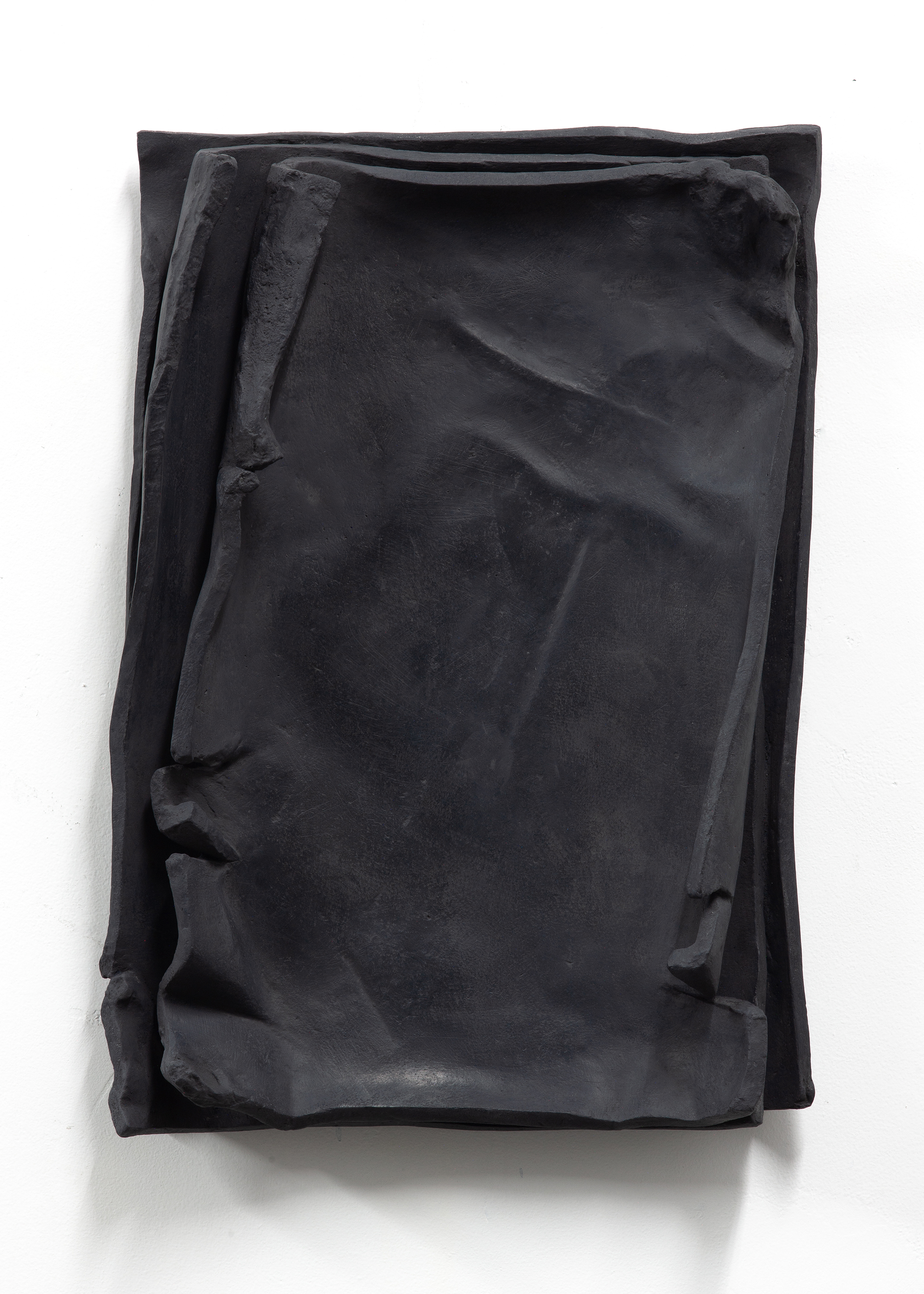 Front view of a matte black relief sculpture by sculptor Erik Andersen. The work is displayed vertically on a white wall. The artwork appears to consist of several loosely stacked sheets resembling paper. However, the epoxy resin sculpture is made from a single piece.