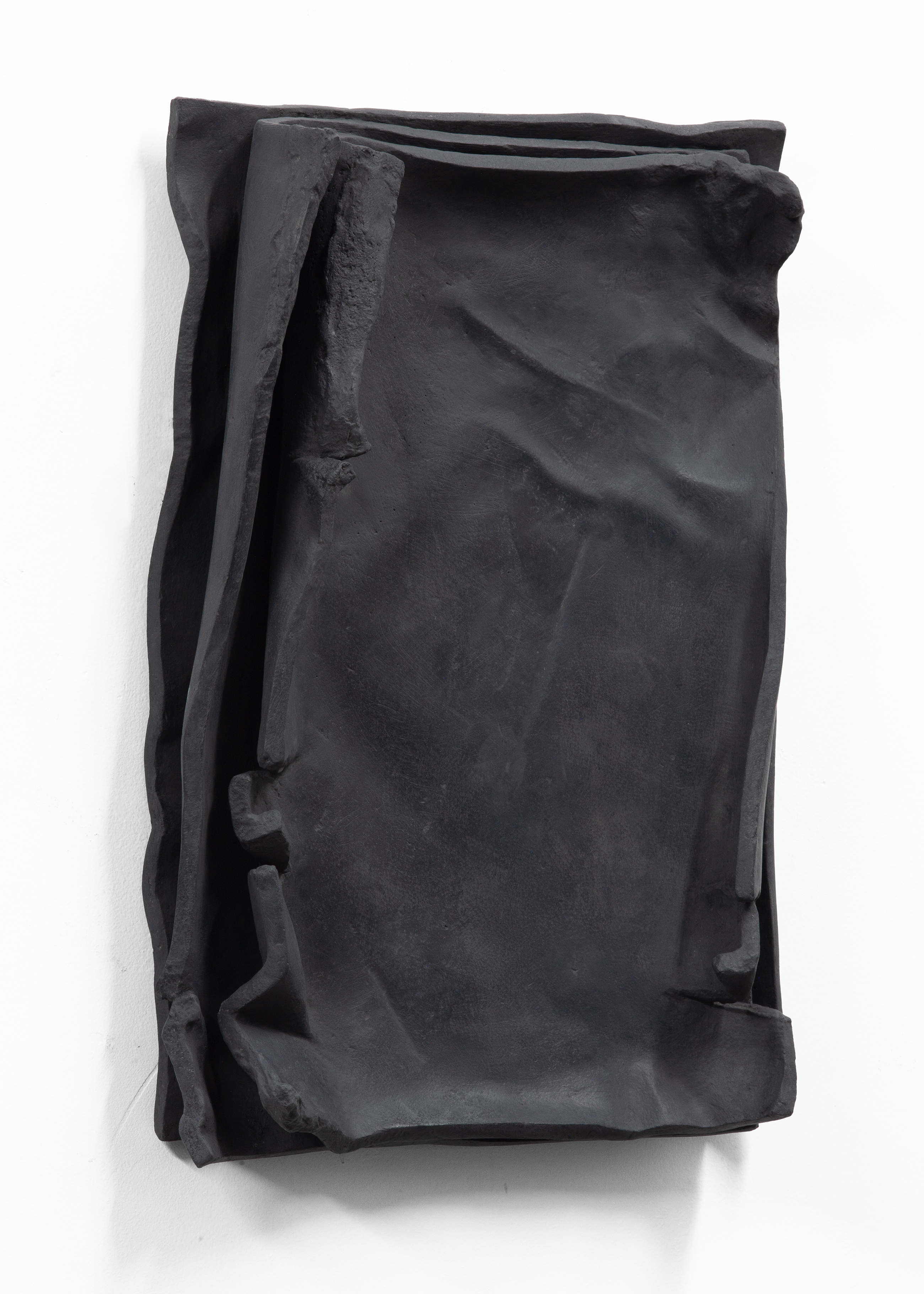 Side view of a matte black relief work titled Besser Vertikal 12 by sculptor Erik Andersen. In keeping with the title, the artwork is displayed vertically on a white wall. The work is a solid piece, even though it appears to consist of several sheets layered on top of each other, with curled edges and partially destroyed corners.