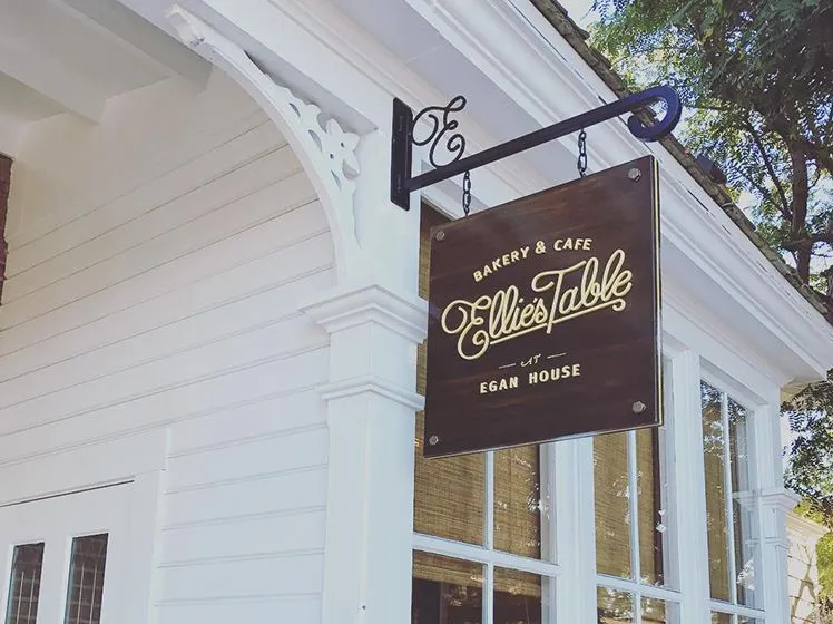 Wooden hanging sign for Ellie's Table Bakery & Cafe at Egan House on white building exterior with windows and decorative trim.