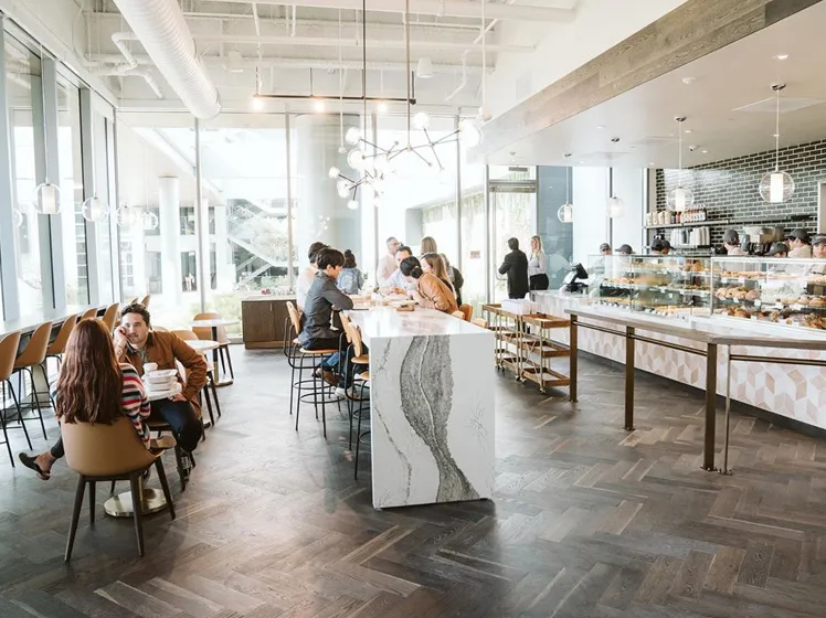 Bright modern café interior with people sitting at tables and a central marble bar counter, large windows letting in natural light.