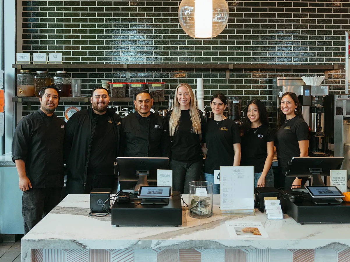 Seven smiling coffee shop employees standing behind a marble counter with cash registers and menu displays.