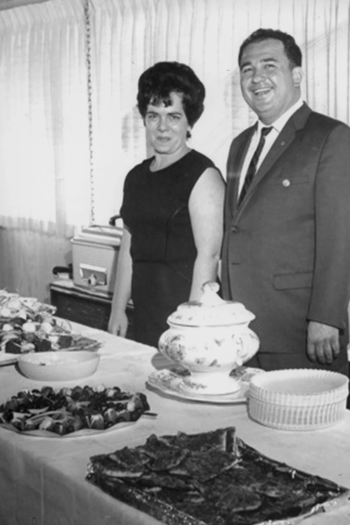 Eleanor and Jay Mastroianni standing together behind a table of food in a family archive photograph.