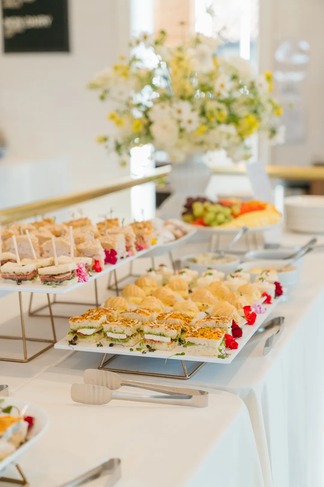Tiered trays of tea sandwiches with fruit and flowers behind them