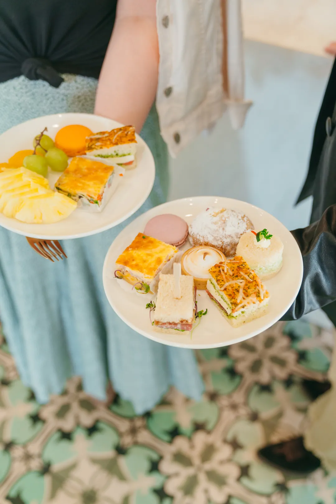 Two guests holding plates of fruit, sandwiches, quiche, tarts and macarons