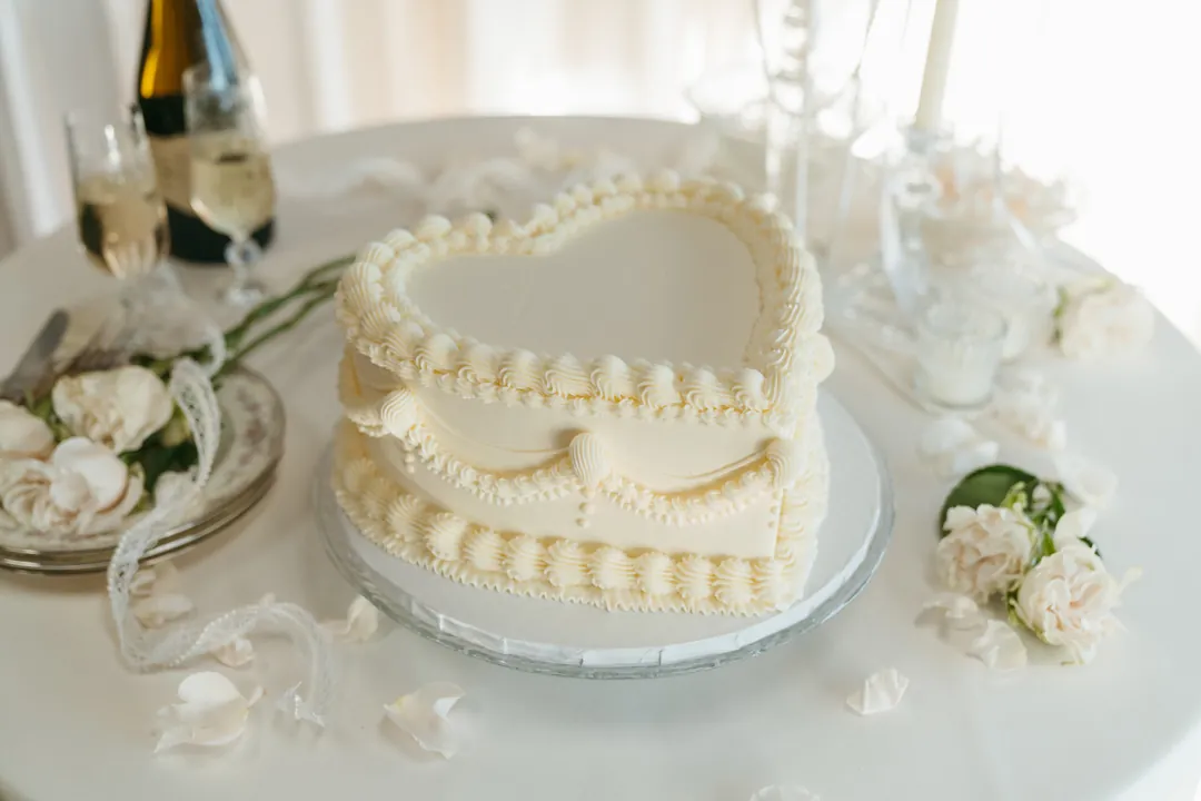 Ivory heart-shaped cake on a white table with champagne and flowers