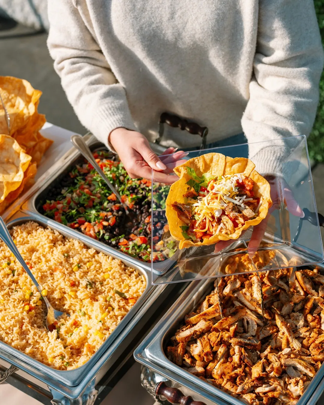 Taco salad bowl held above trays of grilled chicken, rice and black bean salsa