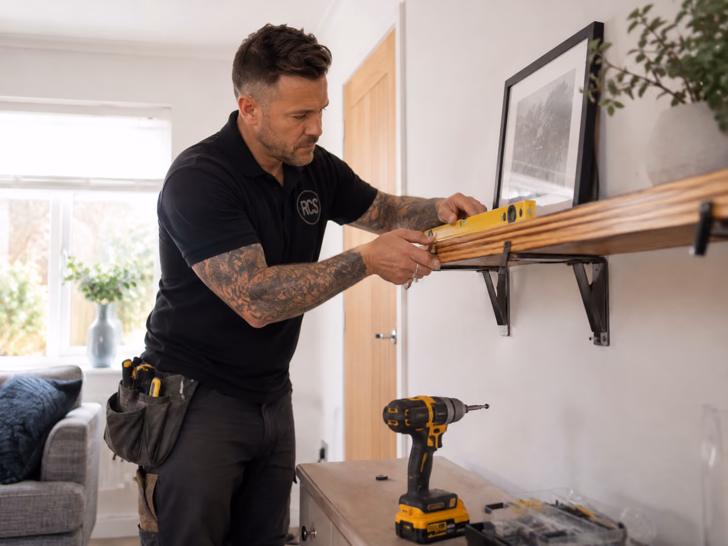Rose Construction Sussex handyman fitting a wooden shelf inside a customer’s home, using a spirit level and drill in a bright living room.