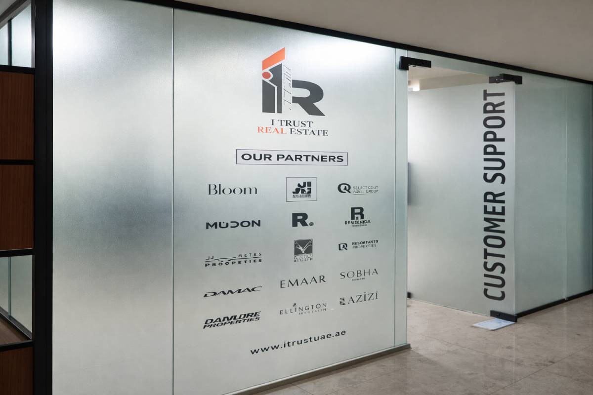 Side eyelevel shot of I Trust Real Estate Frosted Branding Sticker featuring partner logos on glass office walls at Al Darwish Tower, Muroor St, Abu Dhabi, installed by Ninety Nine Advertising LLC.
