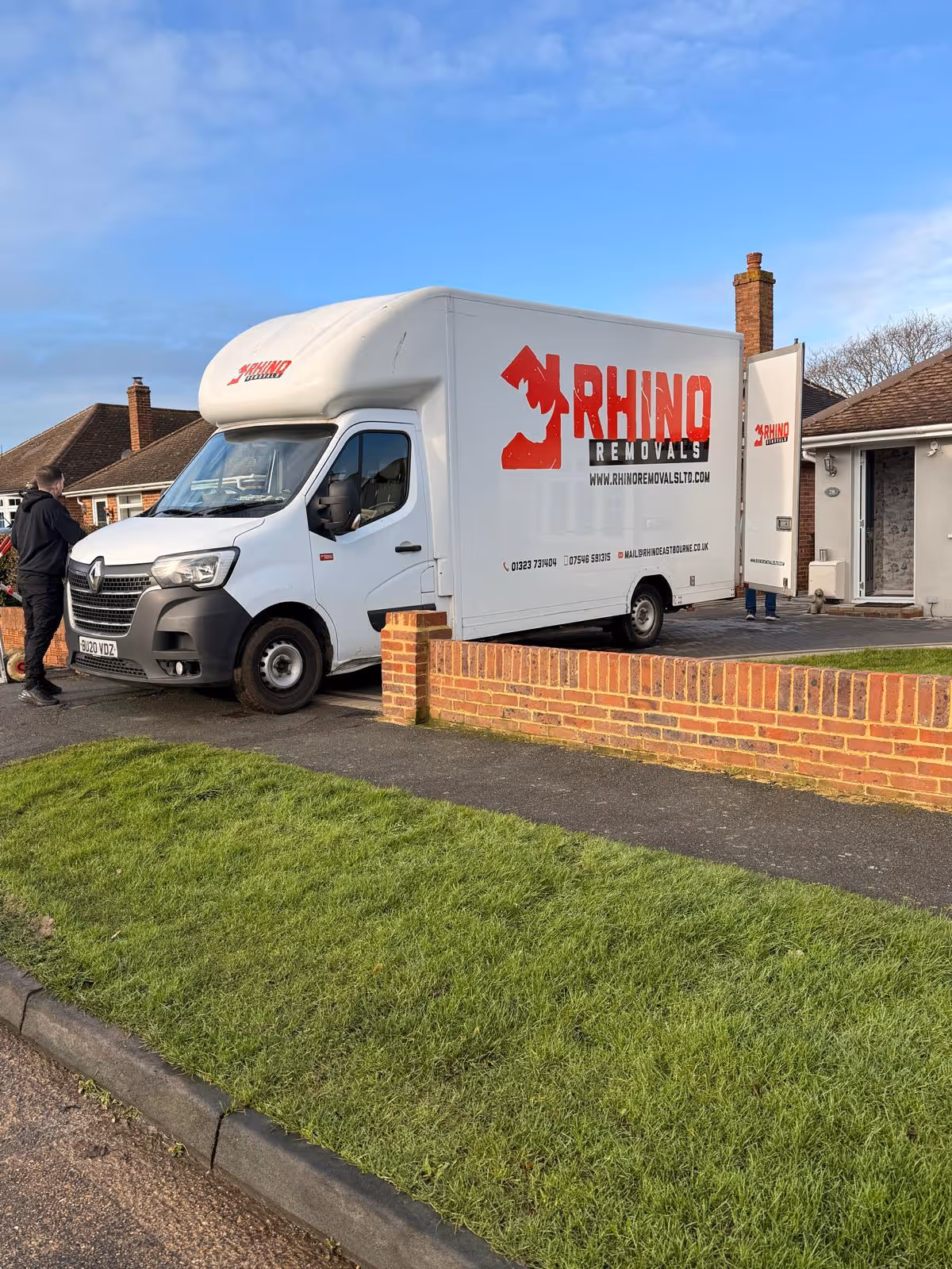 A Rhino Removals truck is parked next to a driveway with a person standing nearby.