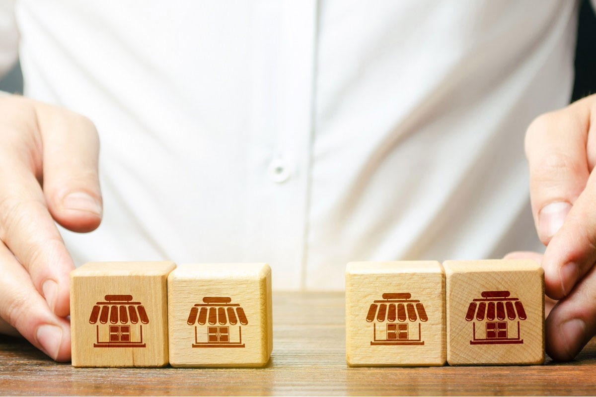 Hands aligning wooden blocks with storefront icons, representing multi-location acquisitions in a franchise-style roll-up strategy