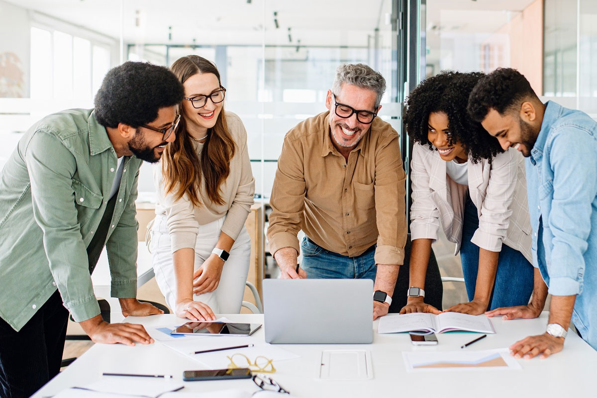 A cheerful and diverse corporate development team gathered around a laptop in a bright, modern office