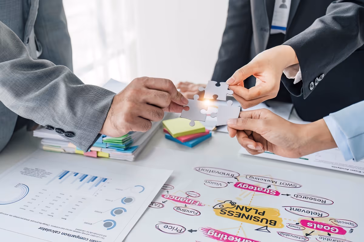 Business professionals in formal attire fitting puzzle pieces together over a desk filled with charts, sticky notes, and a business plan, symbolizing strategic planning and collaboration