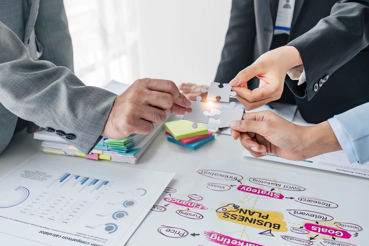 Business professionals in formal attire fitting puzzle pieces together over a desk filled with charts, sticky notes, and a business plan, symbolizing strategic planning and collaboration
