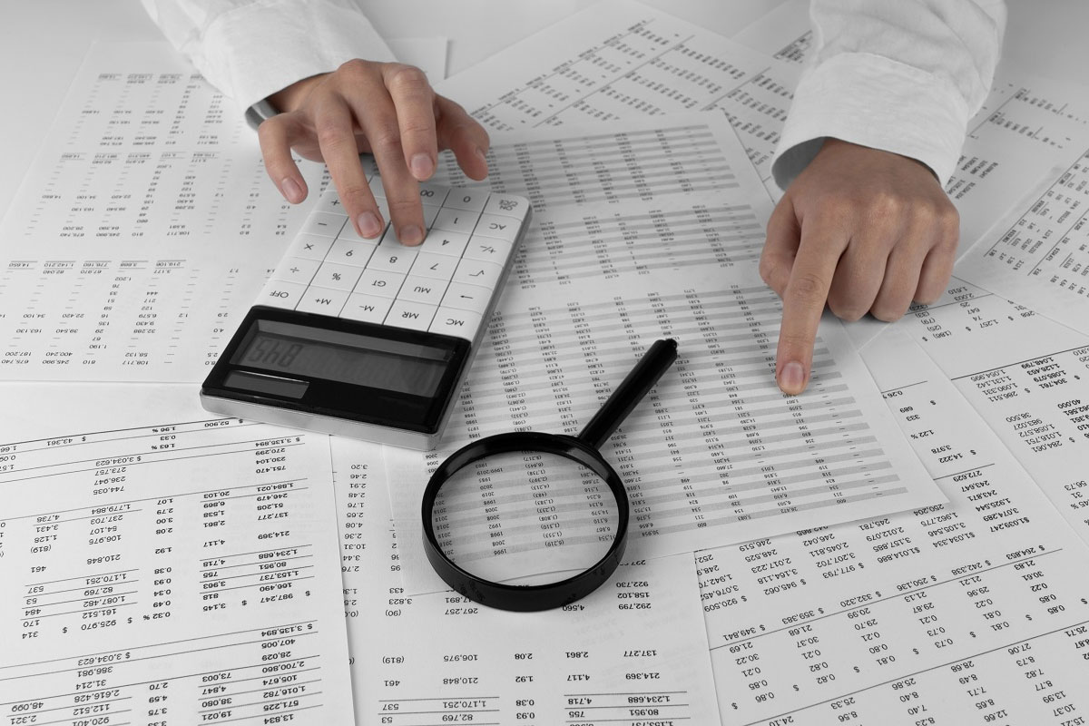 Hands using a calculator and pointing at spreadsheets with a magnifying glass, illustrating financial due diligence and document verification