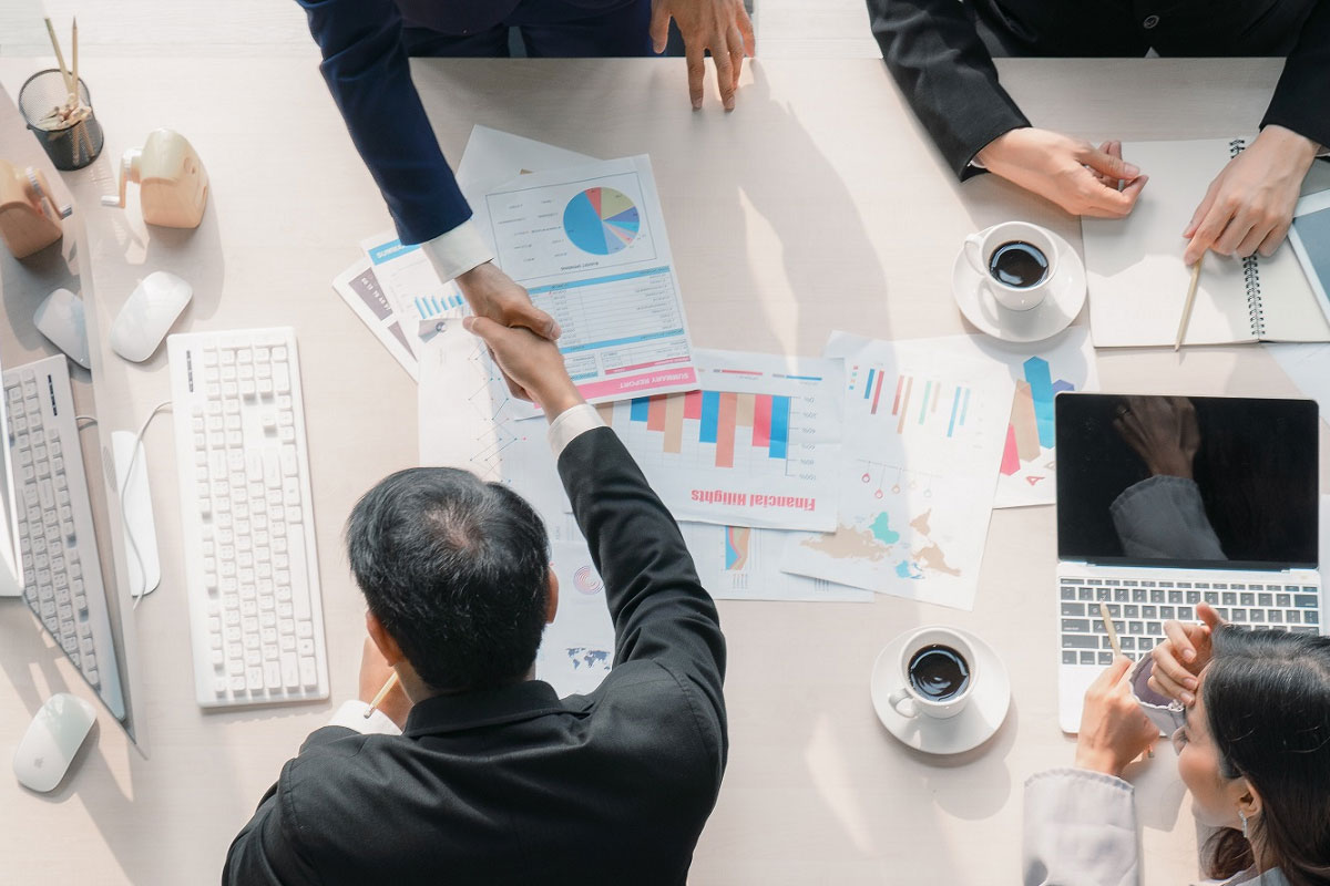 Executives finalizing an M&A deal, using M&A technology solutions around a desk covered with financial charts, analytics reports, and laptops to represent data-driven mergers and acquisitions