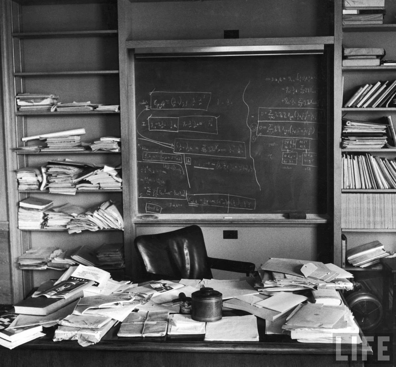 Photographie en noir et blanc d’un bureau couvert de papiers devant un tableau.